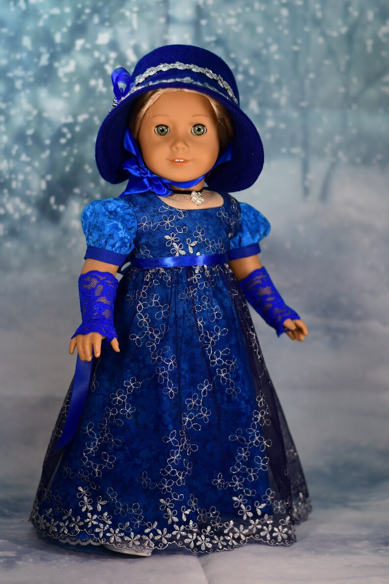 May include: A blue and silver doll's dress with a matching blue hat and gloves. The dress has a floral pattern and a satin sash. The doll is wearing a silver necklace.