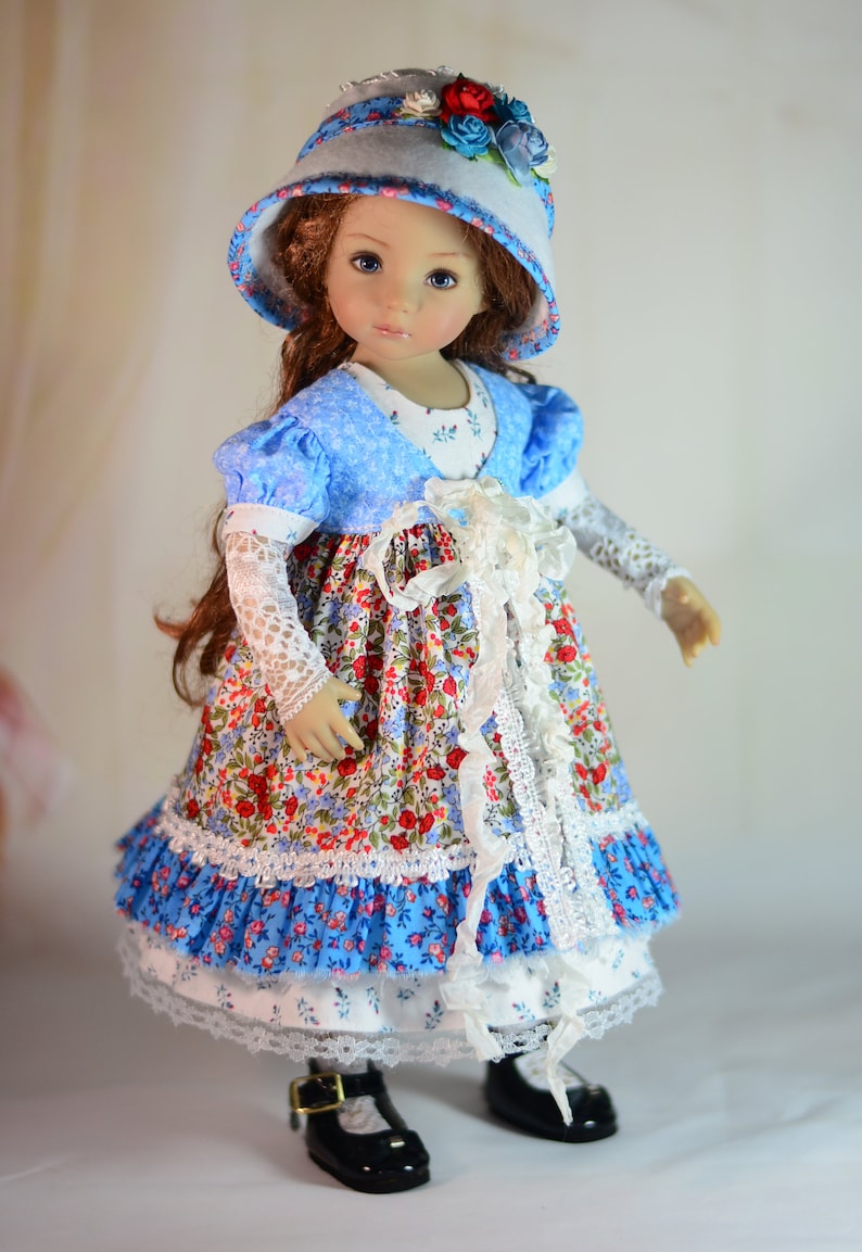 K&ouml;nnte beinhalten: Puppe in einem Kleid mit Blumenmuster, blauem Oberteil und passendem Hut. Das Kleid hat eine wei&szlig;e Spitzenborte und der Hut ist mit Blumen verziert. Die Puppe tr&auml;gt schwarze Schuhe.