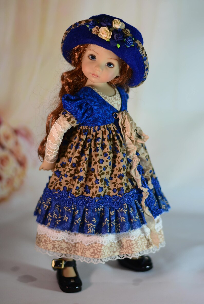 K&ouml;nnte beinhalten: Eine Puppe mit einem blauen Hut, der mit Stoffblumen verziert ist, und einem passenden Kleid. Das Kleid hat ein Blumenmuster mit blauen Akzenten, Spitzenbesatz und einem ger&uuml;schten Rock. Die Puppe hat lange, rotbraune Haare und tr&auml;gt schwarze Schuhe.