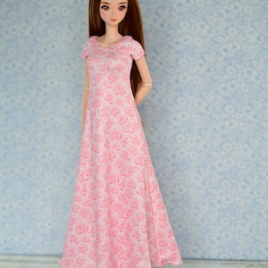May include: A doll wearing a pink dress with a floral pattern and white shoes. The doll has long brown hair and is standing on a light wood floor.