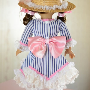 May include: A doll wearing a blue and white striped dress with a pink bow and white lace trim. The doll is also wearing a straw hat with a pink ribbon and white lace trim.