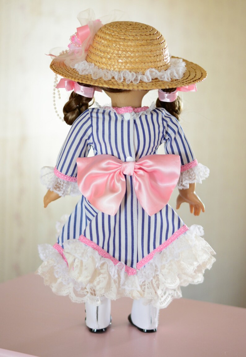 May include: A doll wearing a blue and white striped dress with a pink bow and white lace trim. The doll is also wearing a straw hat with a pink ribbon and white lace trim.