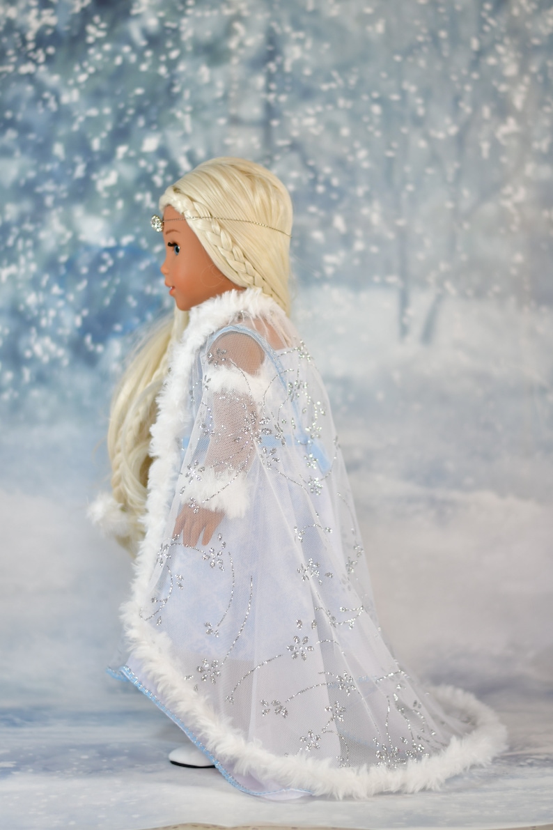 May include: A doll wearing a white and blue dress with a white fur trim and a silver snowflake design. The doll has blonde hair and is wearing a silver crown.