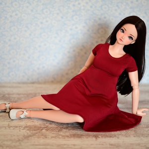 May include: A fashion doll with long black hair and a red dress, seated on a wooden surface. The doll wears white shoes with bows. The background features a light blue wall with a floral pattern.