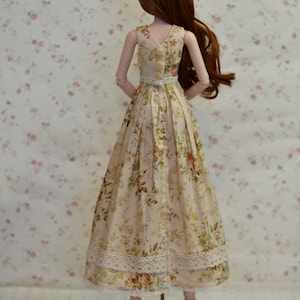 May include: A doll wearing a floral print dress with a white lace trim. The dress has a V-neck and buttons down the back. The doll has long auburn hair and is standing on a wooden base. The dress is cream-colored with a floral pattern in shades of pink, green, and orange.