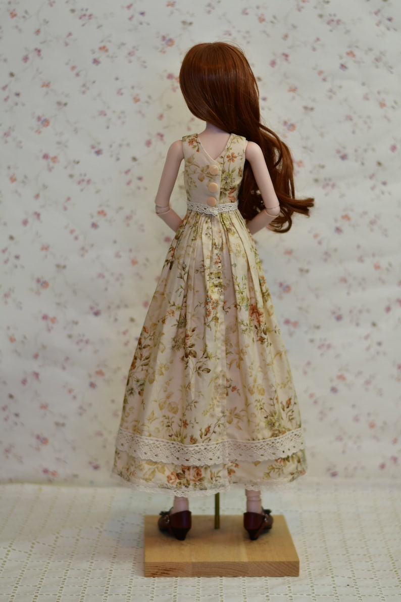 May include: A doll wearing a floral print dress with a white lace trim. The dress has a V-neck and buttons down the back. The doll has long auburn hair and is standing on a wooden base. The dress is cream-colored with a floral pattern in shades of pink, green, and orange.