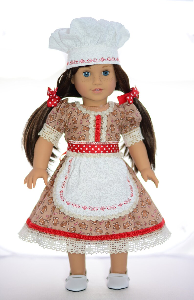 May include: A doll wearing a white chef hat, a brown dress with gingerbread men and candy canes, a red and white polka dot ribbon, and a white apron with red embroidery. The doll has brown hair in pigtails with red polka dot bows.