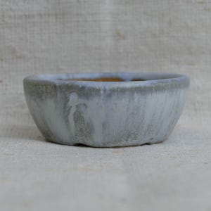 May include: A small, handmade ceramic bowl with a light grey, textured exterior and a brown interior. The bowl has a rounded shape and is suitable for plants or small items.