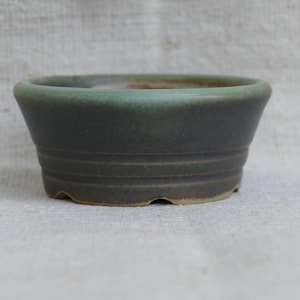 May include: A small, dark green ceramic bonsai pot with a slightly flared shape. The pot has a textured surface with horizontal grooves around the middle and a lighter green rim. The pot sits on a neutral background.