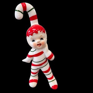 May include: A ceramic Christmas figurine in the shape of a smiling child dressed in a candy cane outfit. The figurine is white with red and green stripes and accents. The head is shaped like a candy cane.