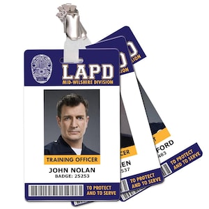 May include: An LAPD Mid-Wilshire Division badge featuring a photo of John Nolan, badge number 25253. The badge is white with blue accents and the text "TO PROTECT AND TO SERVE". A metal clip and lanyard are attached.