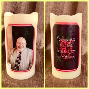 May include: Two white pillar candles with a red border around a photo on one and a red border around a quote on the other. The photo is of a man with a white beard and a checkered tie. The quote reads "You'll always be in my heart, because in there you're still alive."