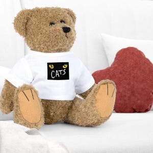 Peut inclure: Un ours en peluche brun portant un t-shirt blanc avec un carré noir et des yeux de chat jaunes qui dit "CATS" dessus.