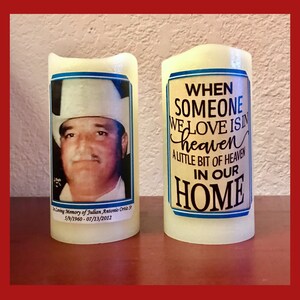 May include: Two white pillar candles with printed labels. One candle has a photo of a man wearing a cowboy hat and the text "In Loving Memory of Julian Antonio Ortiz Sr. 5/9/1960 - 07/13/2012". The other candle has the text "When someone we love is in heaven a little bit of heaven in our home".