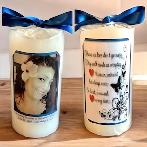 May include: Two white pillar candles with blue ribbon bows. One candle has a photo of a woman with a flower in her hair. The other candle has a black and white design with the text "Those we love don't go away. They walk beside us everyday. Unseen, unheard, but always near. So loved, so missed, so very dear."
