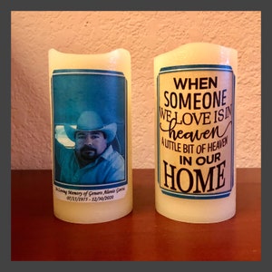 May include: Two white pillar candles with printed labels. One candle has a photo of a man wearing a cowboy hat. The other candle has the text "When someone we love is in heaven a little bit of heaven in our home".