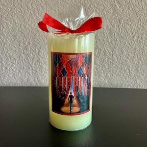 May include: A white pillar candle with a red ribbon tied around it. The candle has a colorful image of a man walking on stage with the word "PIPPIN" printed on it.