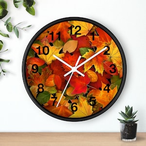 Puede incluir: Reloj de pared redondo con marco negro. La esfera del reloj presenta un collage vibrante de hojas de otoño en tonos rojos, naranjas y amarillos. Las manecillas blancas y los números negros marcan la hora. Una pieza decorativa para el hogar o la oficina.