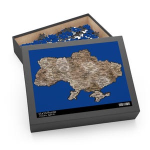 Puede incluir: Una caja de rompecabezas gris con un fondo azul y un mapa de Ucrania. El rompecabezas mide 28x36 cm y tiene 252 piezas. La caja está abierta, revelando algunas piezas del rompecabezas.