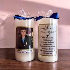 May include: Two white pillar candles with gold trim and a photo of a man in a uniform. The text on the candle reads "In Loving Memory of Grand Bill October 27, 1938 - May 17, 2015" and "until we meet again".