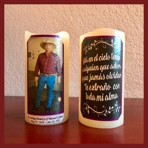 May include: Two white pillar candles with printed labels. The left candle has a photo of a man wearing a plaid shirt and a cowboy hat. The label reads "In Loving Memory of Manuel Gomes May 17, 1954 - Jan 19, 2021". The right candle has a black label with white text that reads "Alla en el cielo tengo alguien que adorn yque jamás olvidar Te extraño con toda mi alma."