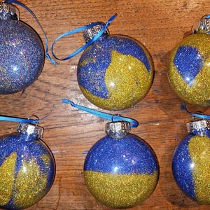 May include: Six glittery Christmas ornaments. Each glass ball is decorated with blue and gold glitter, with a silver cap and a blue ribbon for hanging. The ornaments are arranged on a wooden surface.