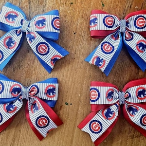 Op de afbeelding: Vier decoratieve haarstrikken met het Chicago Cubs-logo. Twee strikken zijn blauw en wit, en twee zijn rood en wit. Elke strik heeft een zilveren glittercentrum en een gestreept patroon.