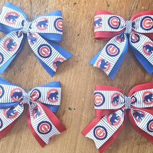 May include: Four decorative hair bows. Two are blue and white with red accents, and two are red and white with blue accents. Each bow features a repeating pattern of a bear and the letters "CUB" inside a circle.