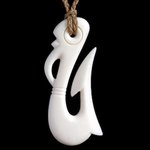 Peut inclure: Collier pendentif de couleur ivoire avec un motif de crochet de pêche stylisé. Le pendentif est suspendu à un cordon tressé beige. Le crochet a une forme incurvée avec un dessus détaillé et une extrémité pointue, sur fond noir.