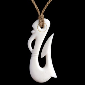 Peut inclure: Un pendentif en os blanc sculpté en forme d'hameçon de pêche traditionnel maori, appelé Hei Matau, suspendu à un collier en corde brune.