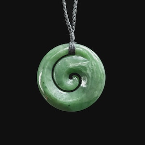 May include: A green jade pendant with a spiral design, suspended from a gray cord. The circular pendant has a polished surface and a dark spiral carved into its center. The background is solid black.