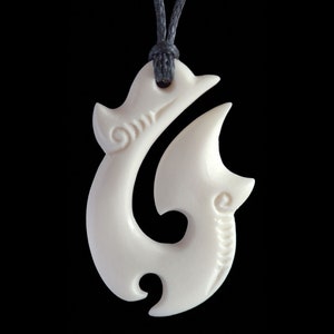 May include: Ivory-colored bone pendant in a stylized fish hook design, suspended from a dark gray cord. The pendant features curved lines and spiral details, set against a black background.