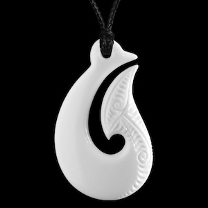 May include: A white pendant necklace with a stylized hook design. The pendant features carved spiral and line patterns and hangs from a black cord. The background is solid black, highlighting the pendant's shape and details.