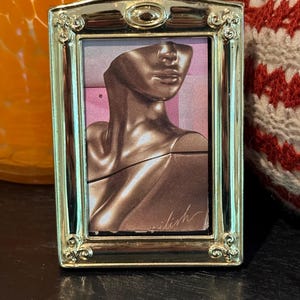 May include: A small, rectangular, silver-colored picture frame. The frame holds a portrait of a person with a brown complexion. The portrait is a collage with the word "eilish" written in cursive. The frame has ornate detailing.
