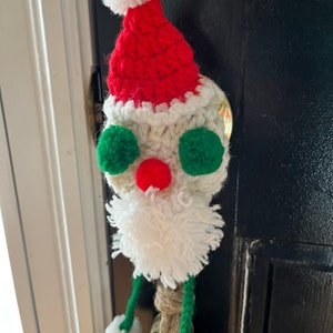 May include: A crocheted Santa Claus ornament with a red hat, white beard, and green eyes. The ornament is attached to a gold bell.