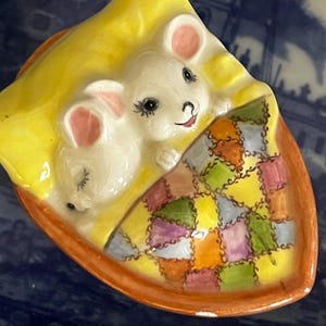 May include: A ceramic figurine of two white mice sleeping in a yellow and brown bed with a colorful patchwork quilt. The mice have pink ears and black eyes.