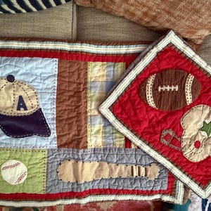 May include: A rectangular quilted fabric piece featuring a baseball cap with the letter A, a baseball, and a bat, alongside a smaller red quilted piece displaying a football and a helmet with a green star, both with decorative stitched borders.