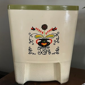 May include: A white ceramic tea canister with a green lid and a floral design. The canister has the word "TEA" written on it.