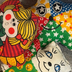 May include: A collection of colorful, handmade fabric toys. The toys include a star with a smiling face and a cat face, and a doll face. The toys are made of various patterned fabrics in red, yellow, green, and blue.