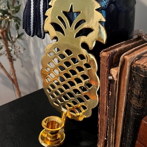 May include: A gold-colored pineapple-shaped candle holder. The candle holder has a detailed, openwork design and a small cup to hold a candle. The pineapple is mounted on a small base. The candle holder is made of metal and has a shiny finish.