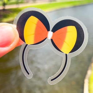 May include: A die-cut sticker featuring a pair of black mouse ears on a thin headband. The ears are accented with a bright orange and yellow candy corn-inspired bow in the centre. The sticker has a clear border and a smooth, glossy finish.