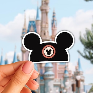 Op de afbeelding: Zwarte Mickey Mouse oren hoed sticker met een witte rand. De hoed heeft een cirkelvormig ontwerp met een Mickey Mouse hoofd in het midden. De achtergrond toont een wazige afbeelding van een kasteel en blauwe lucht.
