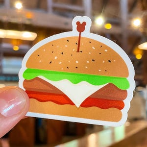 Op de afbeelding: Een kleurrijke sticker in de vorm van een hamburger. De sticker heeft een sesambroodje, sla, tomaat, een patty en kaas. Een kleine tandenstoker in de vorm van een beer is in het bovenste broodje gestoken.