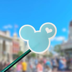 May include: A light blue sticker shaped like a mouse head silhouette with a white outline and a small white heart icon on the right ear. The sticker is attached to a thin, dark green stick, creating a handheld prop against a blurred background.