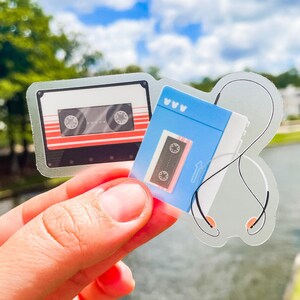 May include: Two die-cut stickers featuring retro audio technology. One sticker depicts a classic black and white cassette tape with red horizontal stripes. The second sticker shows a blue cassette tape connected to a pair of white wired earbuds.