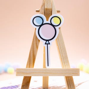 May include: A sticker shaped like a balloon with a circular center and two smaller circular ears in light pink, light blue, and pale yellow, outlined in black. The sticker is displayed on a small wooden easel stand.