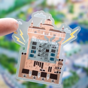 Peut inclure: Un autocollant transparent représentant une illustration beige et marron de The Hollywood Tower Hotel avec des fenêtres bleues, des escaliers de secours noirs et des éclairs jaunes sur les côtés. Le texte The Hollywood Tower Hotel est en blanc.