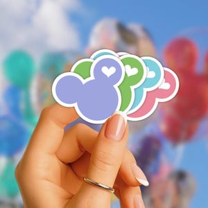 May include: A set of four die-cut stickers shaped like mouse heads, each featuring a small white heart cutout at the top. The stickers are colored in light purple, lime green, light blue, and pink, all outlined with a thick white border.