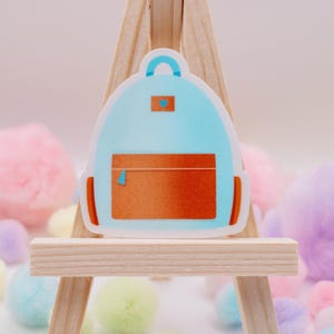 May include: A light blue backpack sticker with a brown pocket and orange accents. The sticker has a small blue handle and a small blue square on the top. The sticker is on a wooden easel with colorful pom-poms in the background.