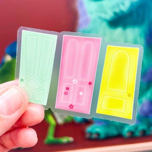 May include: A clear sticker featuring three rectangular doors in light green, pink, and yellow. The green door has geometric paneling, the pink door is decorated with small white and red flower shapes, and the yellow door has a simple arched top.
